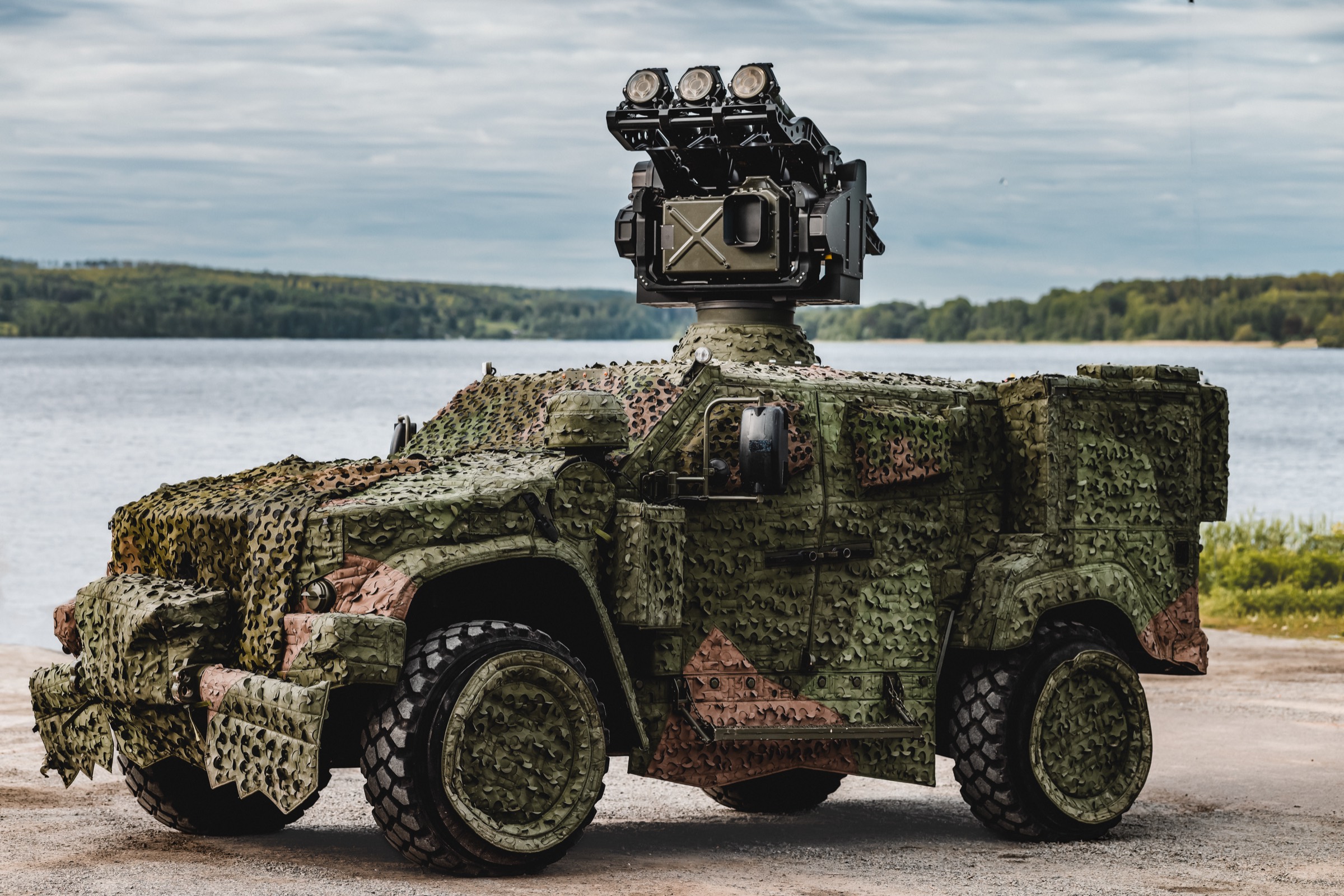 A camouflaged Oshkosh vehicle carrying a mast-mounted RBS 70 NG launcher and sight, parked by a lake